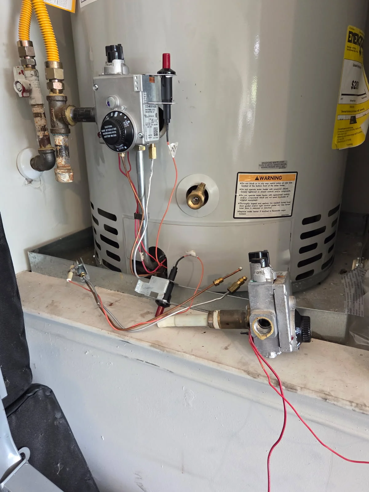 A licensed plumber from Yado's Plumbing inspecting a residential tank-style water heater in a Pasadena home utility closet. The technician checks connections and the pressure relief valve on an aging unit showing signs of sediment buildup.